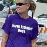 Person wearing Genesis Assistance Dogs shirt at outdoor event.